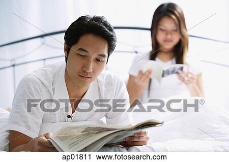 Couple on bed, man reading newspaper, woman behind him reading book View Large Photo Image Stock Image - Couple on bed, man reading newspaper, woman behind him reading book. Fotosearch