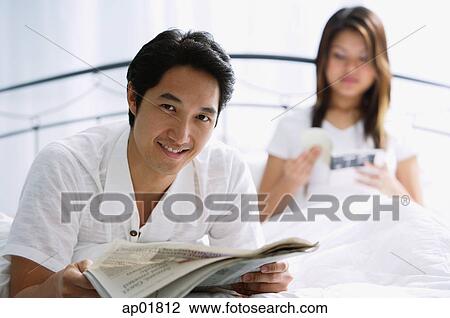 Stock Image - Couple on bed, man with newspaper, smiling, woman behind him reading book. Fotosearch