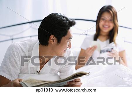 Couple on bed, man with newspaper, smiling at woman behind him View Large Photo Image Stock Image - Couple on bed, man with newspaper, smiling at woman behind him. Fotosearch