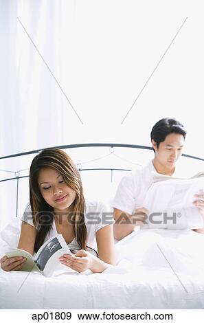 Stock Photo - Couple on bed, woman reading book, man reading newspaper. Fotosearch