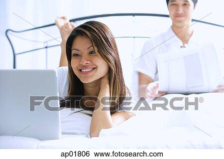 Couple on bed, woman using laptop, man behind her, reading newspaper View Large Photo Image Stock Photograph - Couple on bed, woman using laptop, man behind her, reading newspaper. Fotosearch