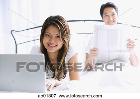 Stock Photo - Couple on bed, woman with laptop, man behind her, reading newspaper, looking at camera. Fotosearch