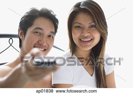Stock Photo - Couple sitting on bed, man holding remote towards camera. Fotosearch