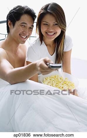 Stock Photograph - Couple sitting on bed, smiling, watching TV. Fotosearch