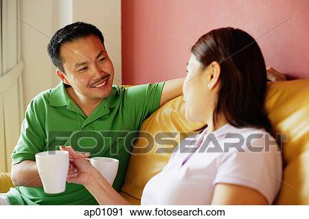 Stock Image - Couple sitting on sofa, side by side, holding mugs. Fotosearch