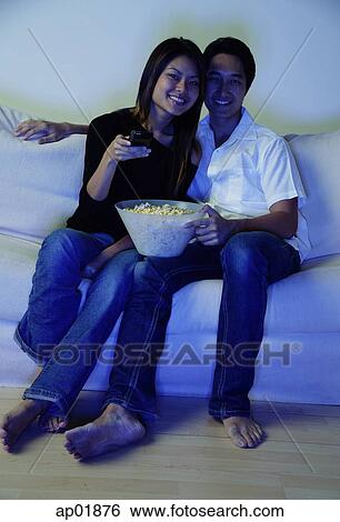 Couple sitting on sofa, watching TV, woman holding remote control View Large Photo Image Stock Photograph - Couple sitting on sofa, watching TV, woman holding remote control. Fotosearch