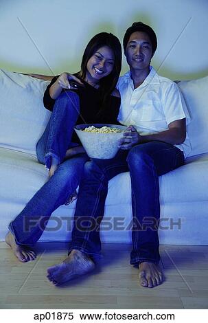 Stock Photography - Couple sitting on sofa, watching TV, bowl of popcorn between them. Fotosearch
