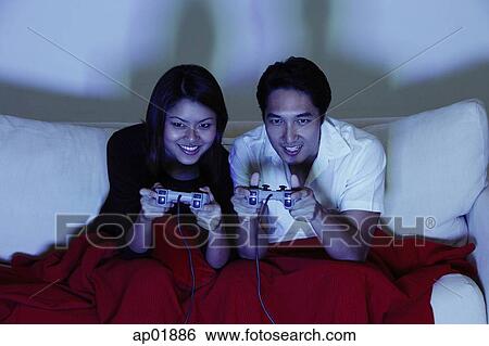 Stock Photograph - Couple sitting side by side on sofa, playing with video games. Fotosearch