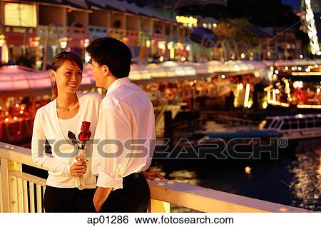 Stock Photograph - Couple standing next to railing, woman holding rose. Fotosearch