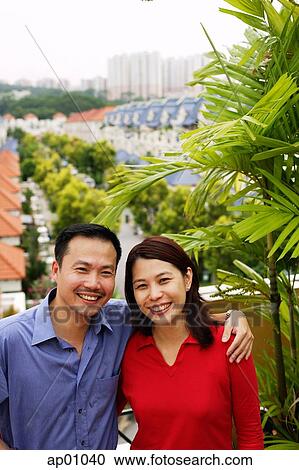 Stock Image - Couple standing on balcony, smiling at camera, portrait. Fotosearch