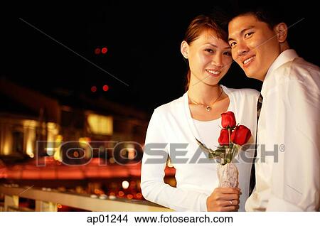 Picture - Couple standing side by side, woman holding single rose stalk, both looking at camera. Fotosearch