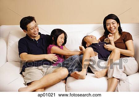 Stock Image - Family of four on sofa, sister tickling brother. Fotosearch