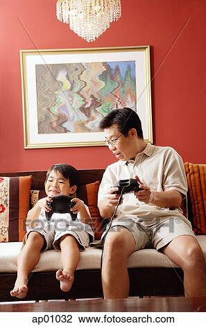 Stock Image - Father and son sitting side by side on sofa, playing video game. Fotosearch