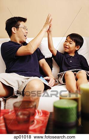 Father and son sitting side by side on sofa, making high five View Large Photo Image Stock Photo - Father and son sitting side by side on sofa, making high five. Fotosearch