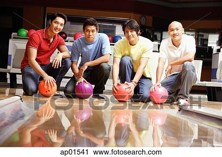 Stock Image - Four men crouching in bowling alley, holding bowling balls. Fotosearch