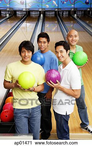 Stock Image - Four men standing in bowling alley, holding bowling balls. Fotosearch