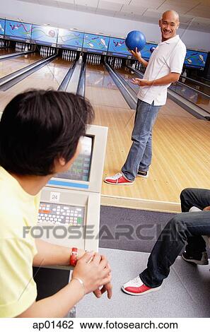 Stock Image - Man holding bowling ball, standing in bowling ally, turning to look at people behind him. Fotosearch