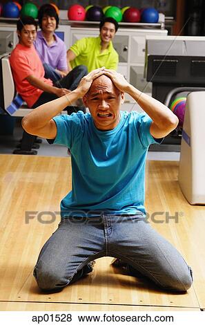 Man kneeling in bowling alley, hands on head, frowning, people in the