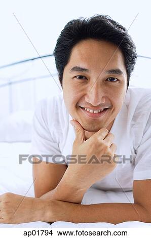 Picture - Man lying on bed, hand on chin, smiling at camera. Fotosearch