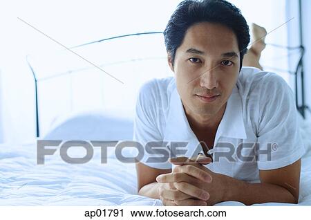Stock Image - Man lying on bed, hands clasped, looking at camera. Fotosearch