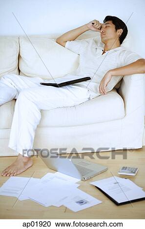 Stock Image - Man lying on sofa, sleeping, hand on head, laptop and documents on floor next to him. Fotosearch