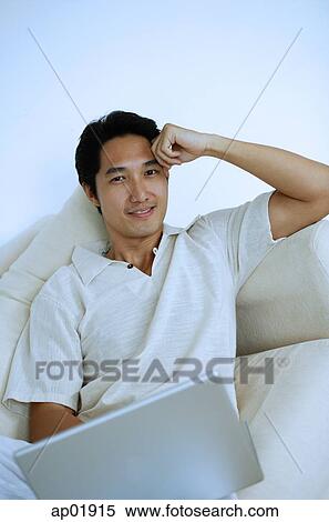 Stock Photography - Man lying on sofa with laptop, hand on head, looking at camera. Fotosearch