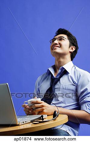 Man sitting in front of laptop, holding mug, looking up View Large Photo Image Picture - Man sitting in front of laptop, holding mug, looking up. Fotosearch