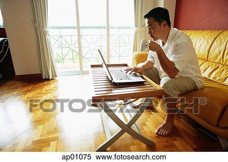 Man sitting in living room, using laptop, drinking from mug View Large Photo Image Stock Photography - Man sitting in living room, using laptop, drinking from mug. Fotosearch
