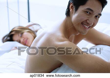 Picture - Man sitting on bed, smiling at camera, woman asleep next to him. Fotosearch