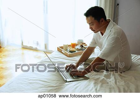 Man sitting on bed, using laptop, breakfast tray next to him View Large Photo Image Stock Photo - Man sitting on bed, using laptop, breakfast tray next to him. Fotosearch