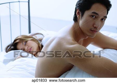 Man sitting on bed, woman asleep next to him View Large Photo Image Stock Image - Man sitting on bed, woman asleep next to him. Fotosearch