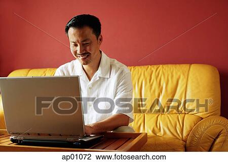 Stock Image - Man sitting on sofa, using laptop. Fotosearch