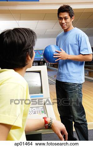 Man standing in bowling ally, holding bowling ball, another man sitting in front of him View Large Photo Image Picture - Man standing in bowling ally, holding bowling ball, another man sitting in front of him. Fotosearch