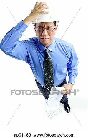 Stock Image - Man wearing hardhat frowning, hand on head, holding roll of paper. Fotosearch