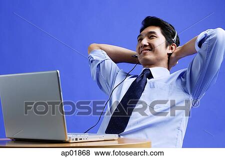 Stock Photo - Man wearing headset, using laptop, leaning back, hands behind head. Fotosearch