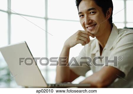 Stock Image - Man with laptop, smiling at camera, hand on chin. Fotosearch
