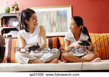 Stock Photography - Mother and daughter on sofa, holding video game controllers, looking at each other. Fotosearch