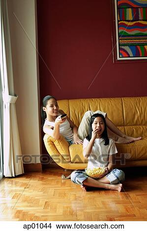 Two girls sitting in living room, one holding remote control, the other eating popcorn View Large Photo Image Picture - Two girls sitting in living room, one holding remote control, the other eating popcorn. Fotosearch