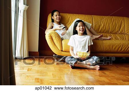 Stock Image - Two girls sitting in living room, one holding remote control, the other holding bowl of popcorn. Fotosearch