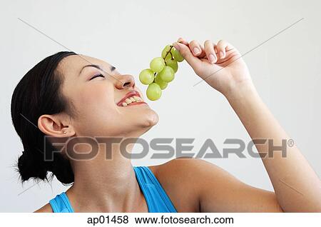 Stock Photo - Woman holding grapes near mouth, side view. Fotosearch