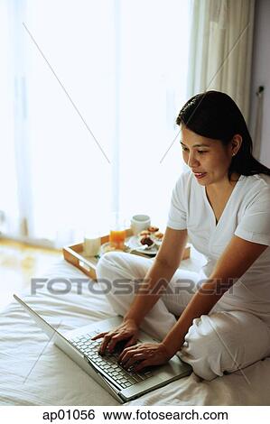 Woman sitting on bed, using laptop View Large Photo Image Stock Photograph - Woman sitting on bed, using laptop. Fotosearch