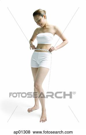 Stock Photo - Young woman standing, holding tape measure around her waist. Fotosearch