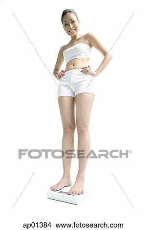 Young woman standing on weighing scale, hands on hips, smiling at camera View Large Photo Image Picture - Young woman standing on weighing scale, hands on hips, smiling at camera. Fotosearch