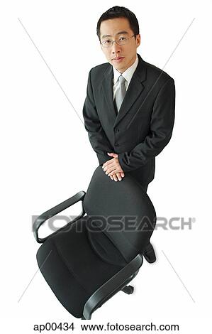 Picture - Businessman standing next to office chair, looking at camera. Fotosearch