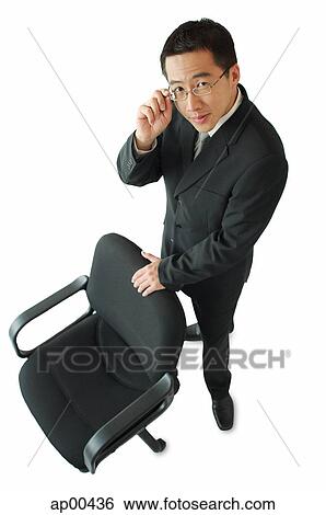 Stock Photograph - Businessman standing next to office chair, adjusting his glasses, looking at camera. Fotosearch