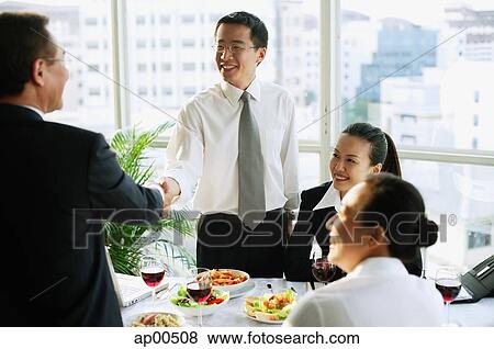 Businessmen shaking hands over lunch table, businesswomen sitting next to them View Large Photo Image Stock Photo - Businessmen shaking hands over lunch table, businesswomen sitting next to them. Fotosearch