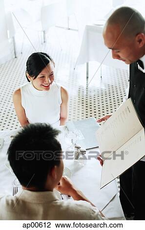 Stock Image - Couple at restaurant, waiter showing them menu. Fotosearch