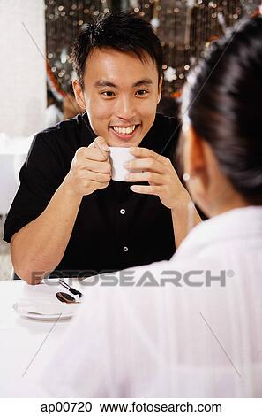 Stock Image - Couple eating in restaurant, man holding cup, over the shoulder view. Fotosearch