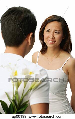 Stock Photo - Couple facing each other, man hiding flowers behind his back. Fotosearch