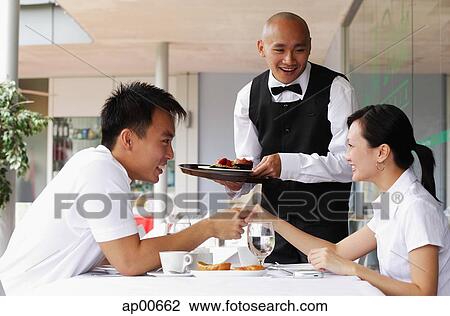 Stock Image - Couple holding hands across the table, waiter standing next to them. Fotosearch
